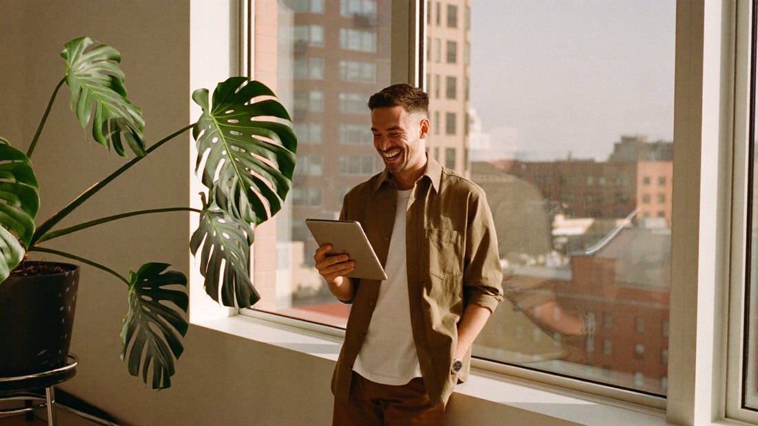 Un indépendant consulte sa tablette en souriant devant la fenêtre de son bureau.